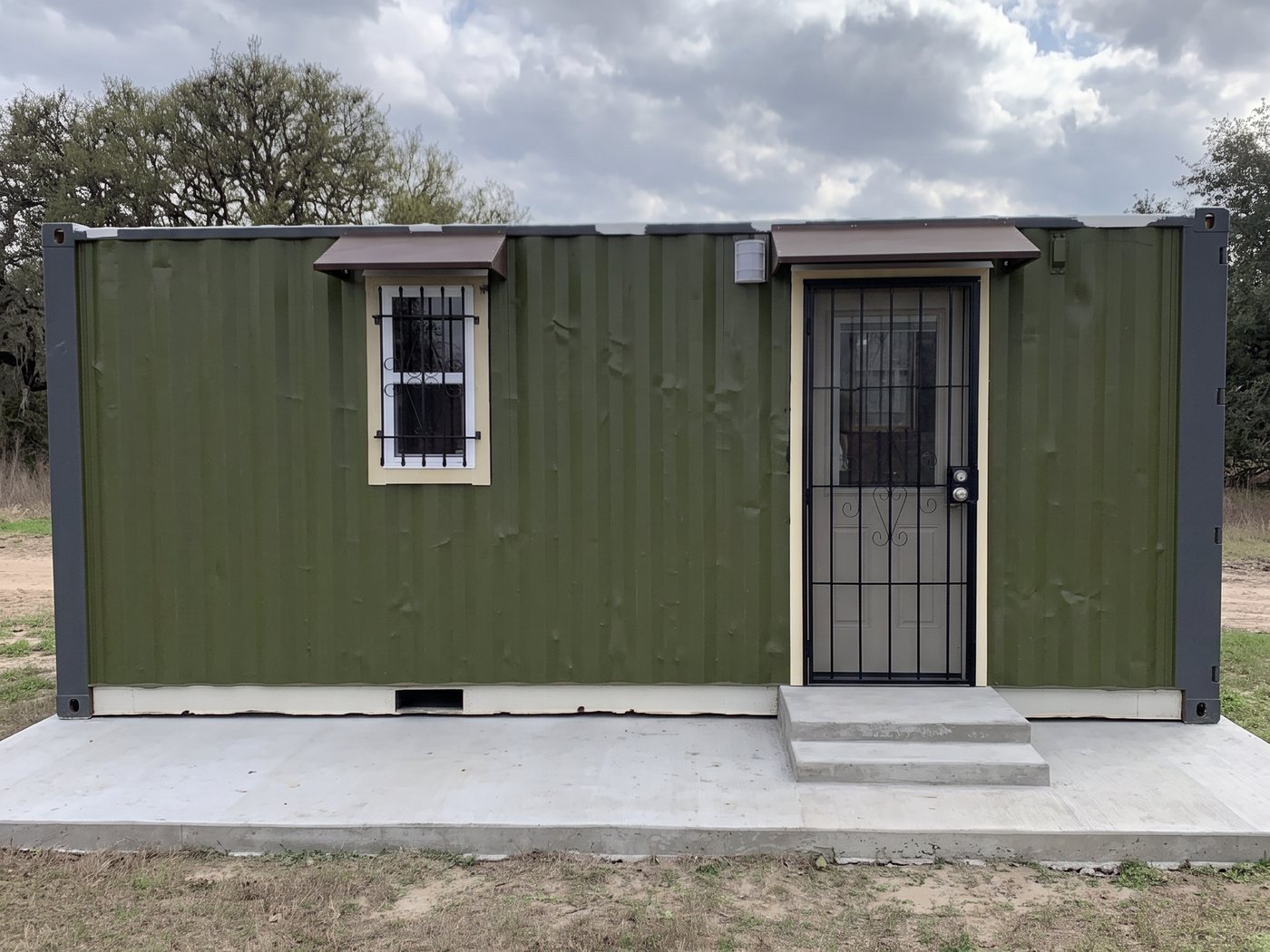 An olive green 20-foot cabin with a security door and barred window on a concrete pad